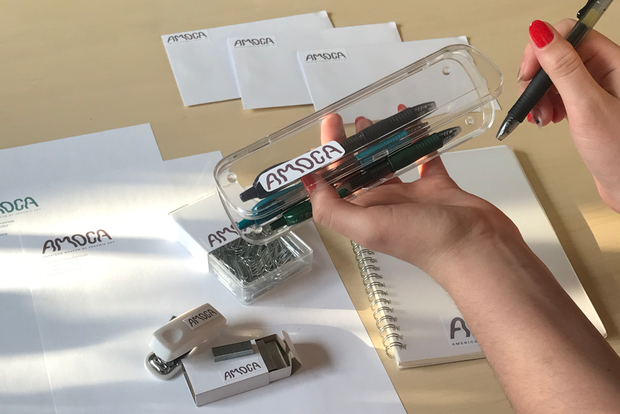 A large image of stationary that was in the pervious image including a letterhead, envelope, and notebook. The pencil case is being held by a hand with red nail polish and another holding a pen
